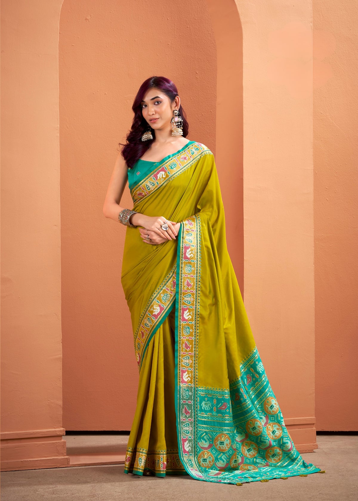 Woman wearing a green saree with a patterned border against a peach-colored wall.