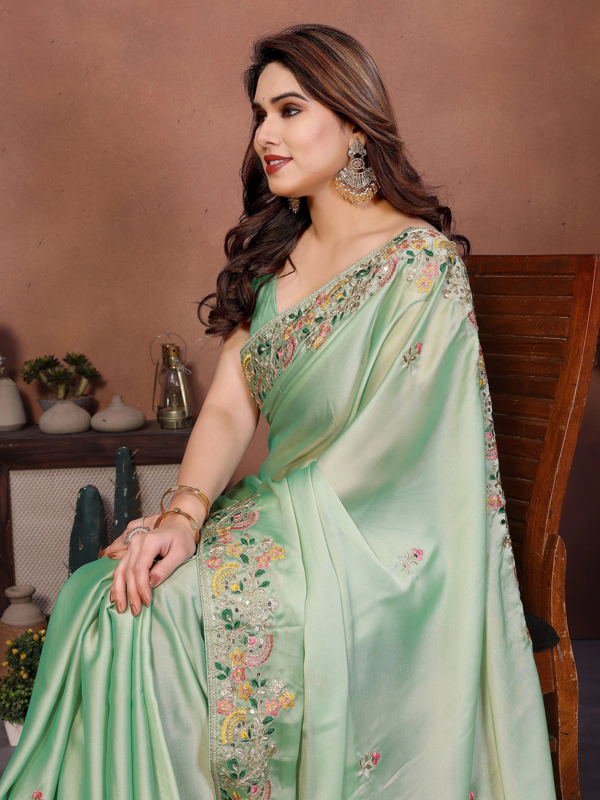 Woman wearing a light green saree with floral embroidery, sitting against a brown background.