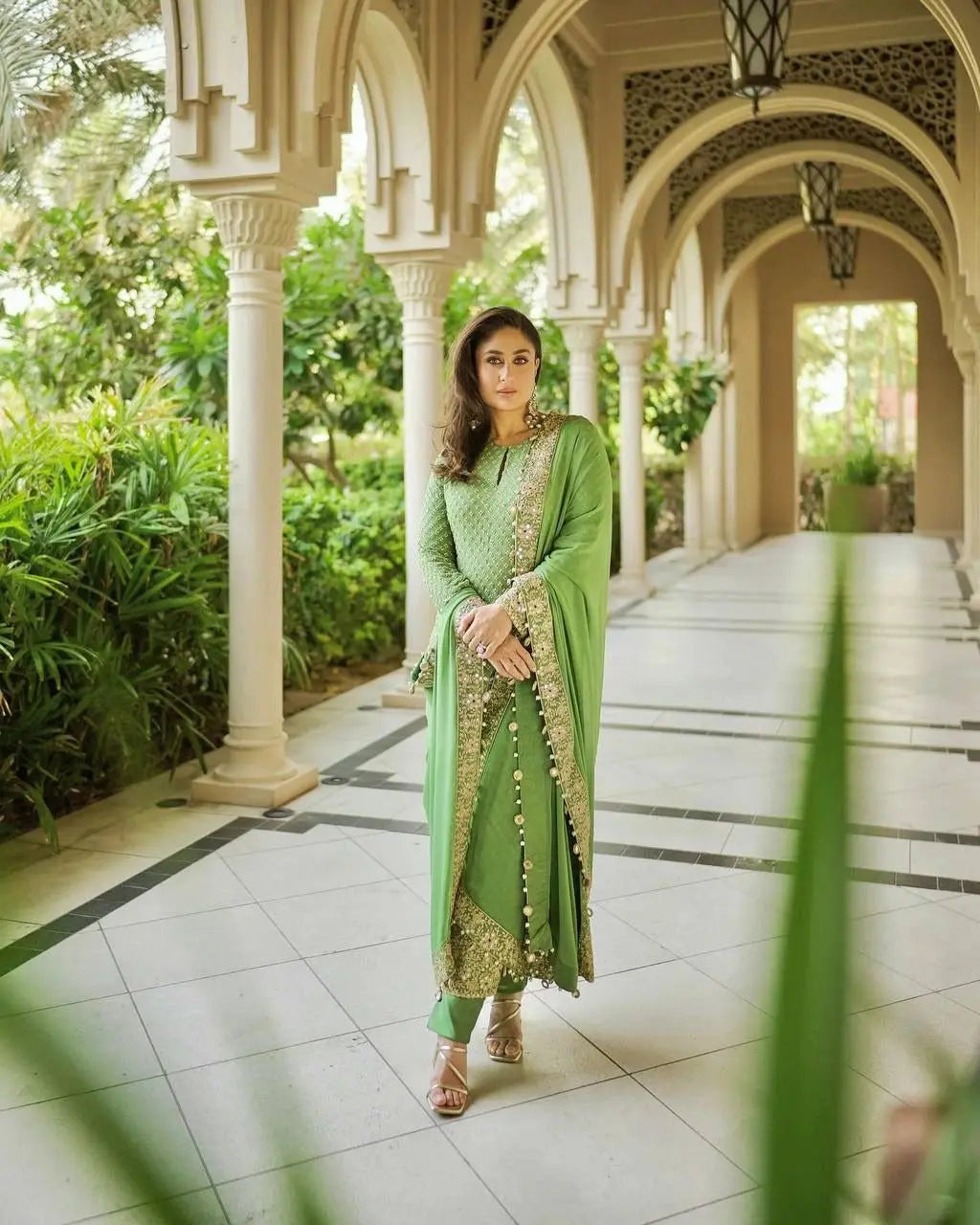 Woman in a green traditional outfit standing in an elegant outdoor setting with arches and greenery.