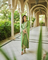Woman in a green traditional outfit standing in an elegant outdoor setting with arches and greenery.