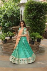 Woman in a green and peach traditional outfit standing outdoors with greenery.
