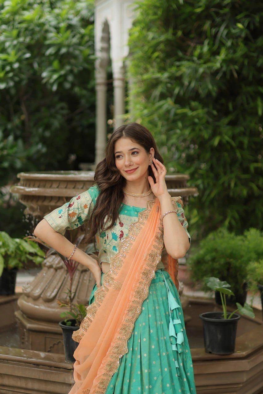 Woman in a green saree with peach border standing outdoors with greenery in the background