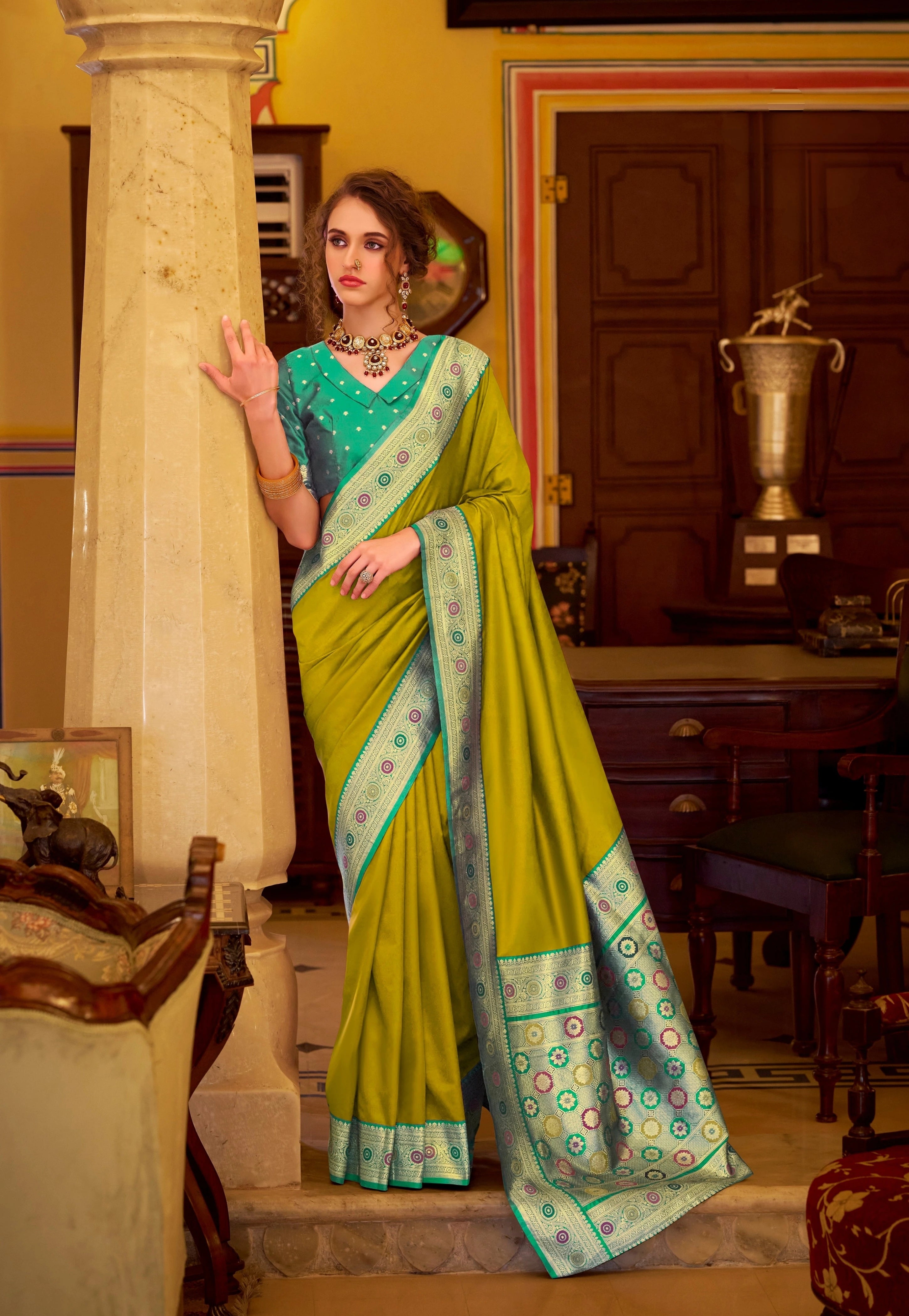 Woman in a green saree with a blue blouse standing in an elegant room.