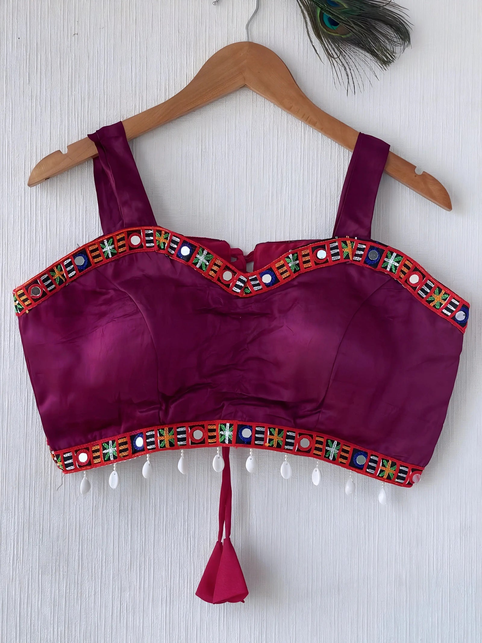 Magenta ethnic blouse featuring vibrant mirror work and a multicolored embroidered geometric border in green, blue, orange, and white, adorned with white pearl drop tassels and a tie-up waist detail. Displayed on a wooden hanger against a textured off-white wall with peacock feather, creating a festive and traditional look.