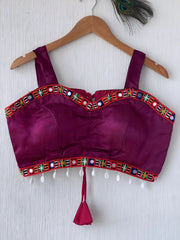  Magenta ethnic blouse featuring vibrant mirror work and a multicolored embroidered geometric border in green, blue, orange, and white, adorned with white pearl drop tassels and a tie-up waist detail. Displayed on a wooden hanger against a textured off-white wall with peacock feather, creating a festive and traditional look.