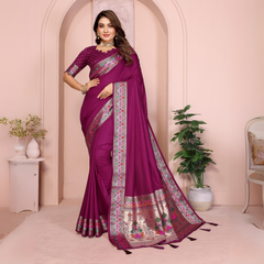 Woman wearing a plum purple silk saree with an ornate multicolor woven border featuring geometric and floral motifs, paired with a matching embroidered blouse and accessorized with a statement choker necklace, bangles, and rings. She poses gracefully in a pastel arched studio decorated with a white side table, floral vase, lush potted green plant, and a vintage-style chair, creating an elegant and sophisticated ambiance.