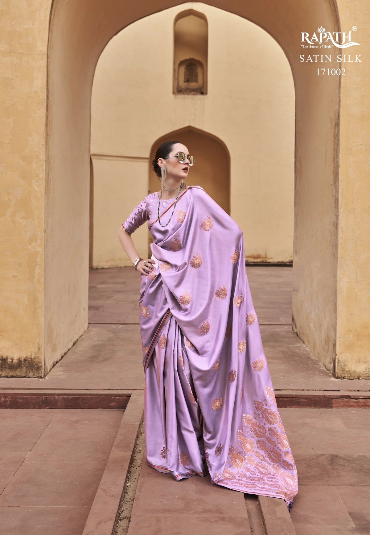 Woman in a lavender saree with gold patterns standing in front of an archway.