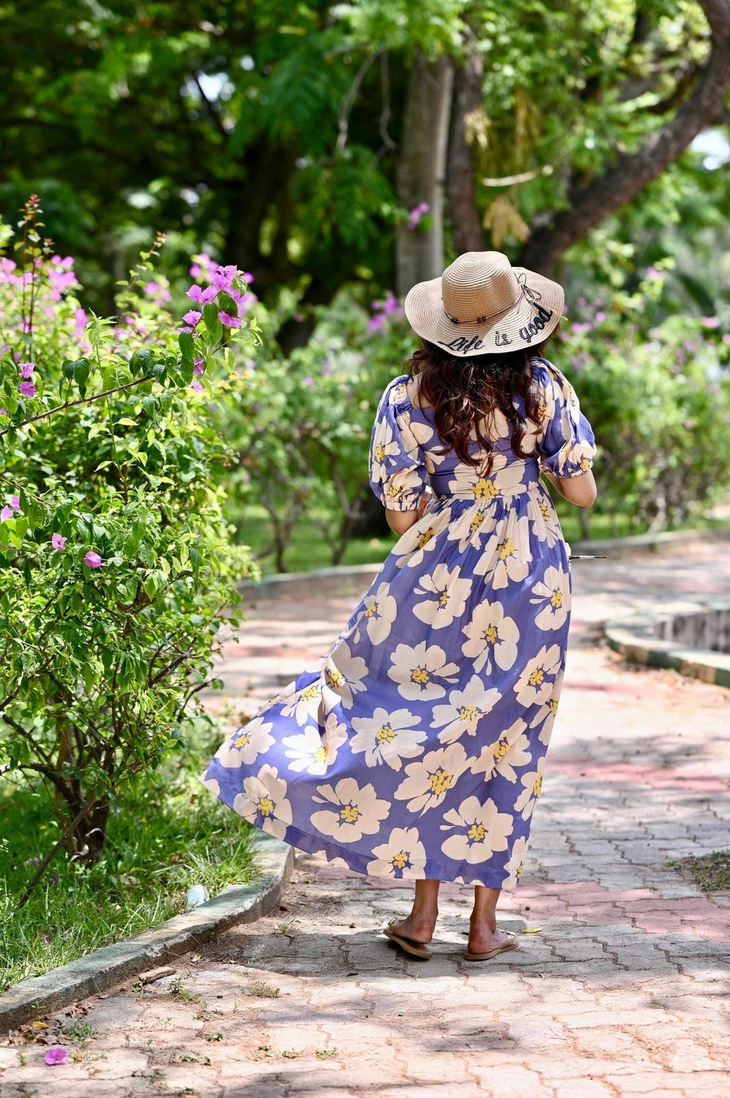 Woman in a blue floral dress and straw hat walking on a garden path.