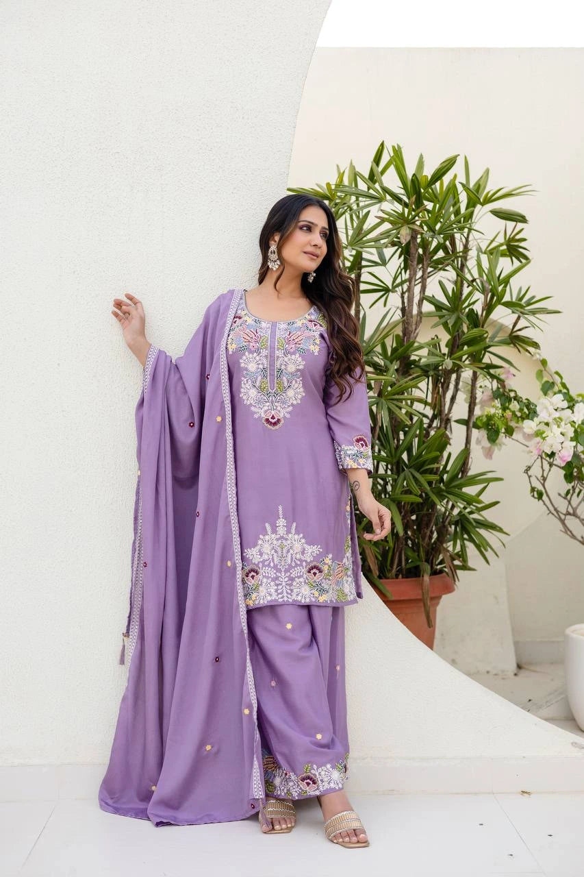 Woman in a lavender traditional outfit with floral embroidery standing next to a plant indoors.