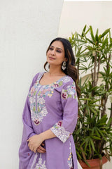 Woman wearing a purple embroidered outfit standing against a white wall with green plants.