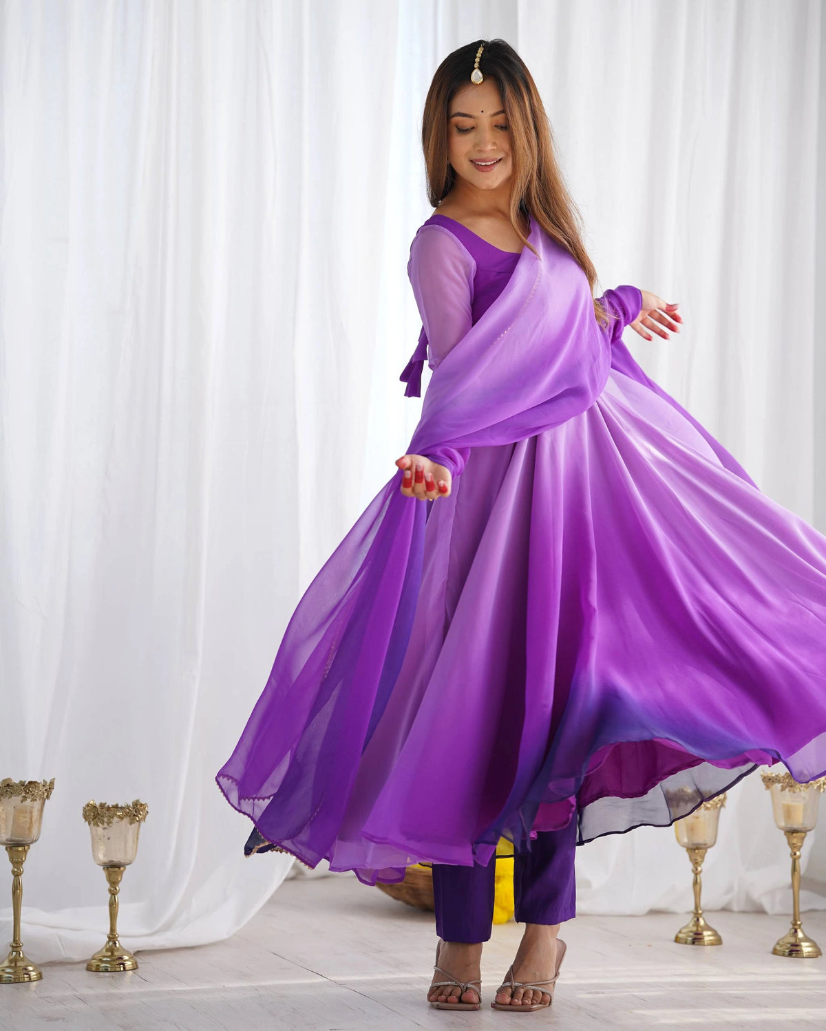 A woman twirls in a long purple ombre Anarkali suit, causing the fabric to flare out, while she looks down with a smile.