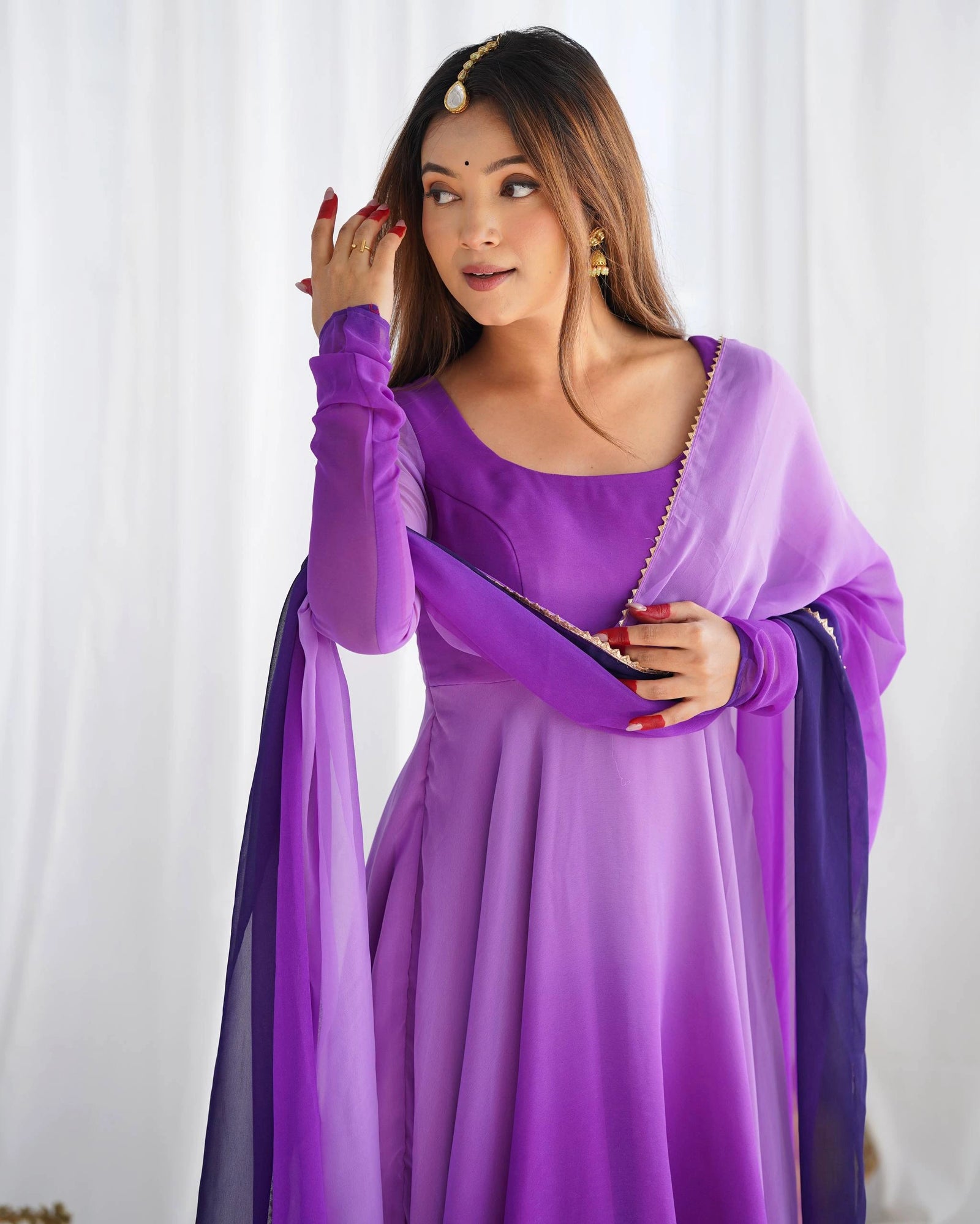 woman sits gracefully on the floor with her purple ombre dress and dupatta spread around her, looking to the side in a bright setting.