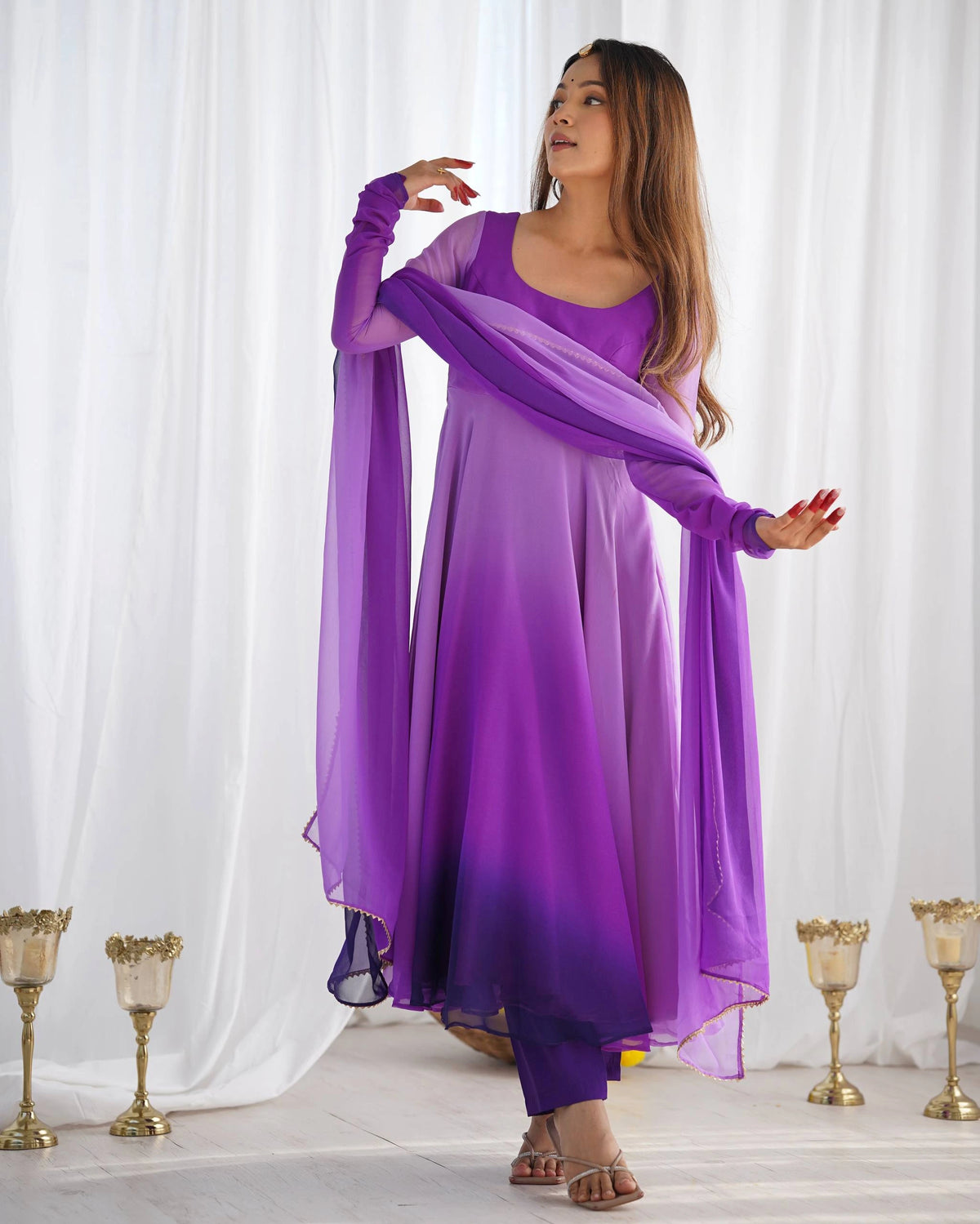 A woman poses standing, looking away with one hand raised, showcasing a purple ombre ethnic outfit with long sleeves and a matching dupatta.