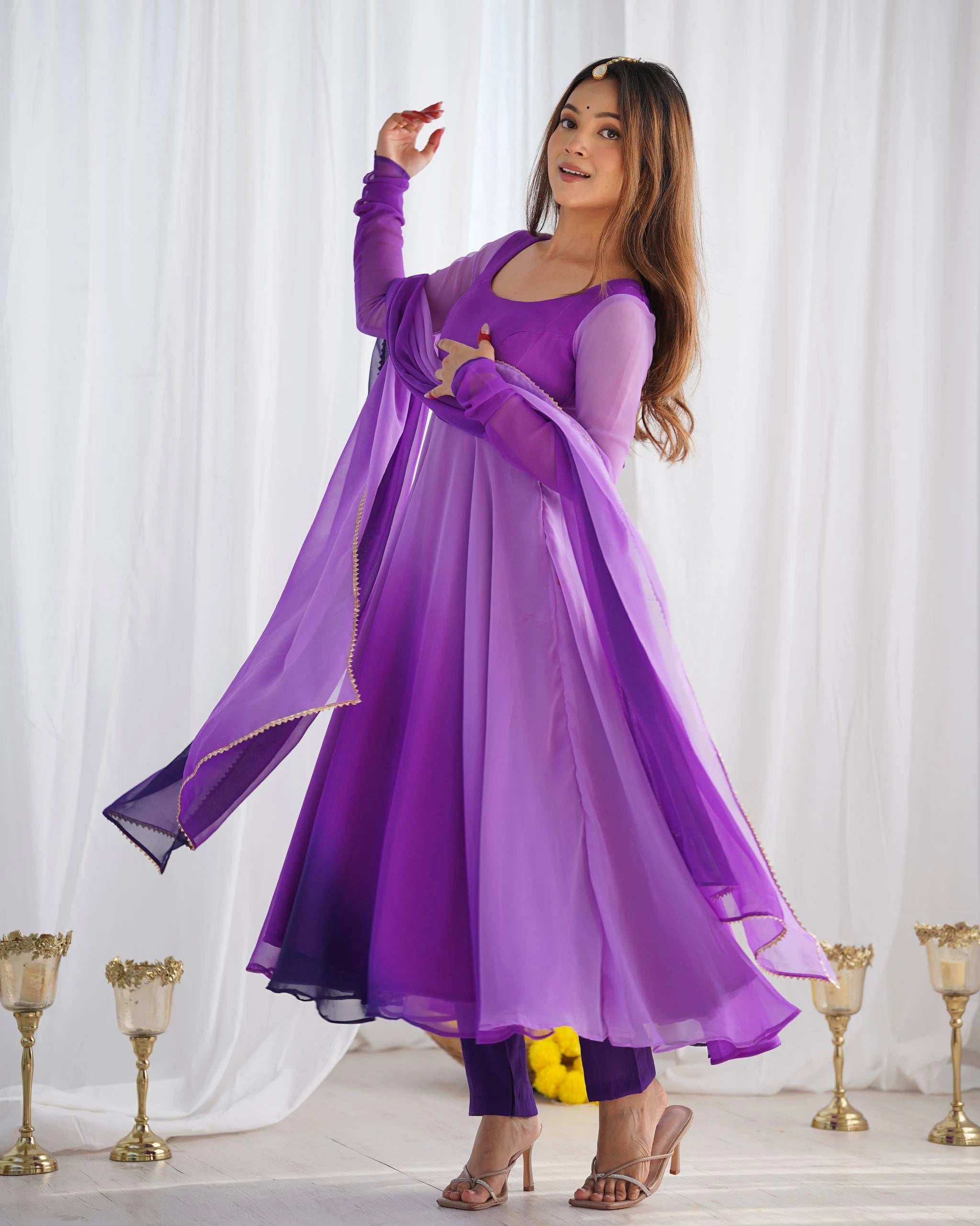 A woman in a flowing purple ombre ethnic dress twirls slightly, holding the edge of her dupatta, accented by a gold "maang tikka" on her forehead.