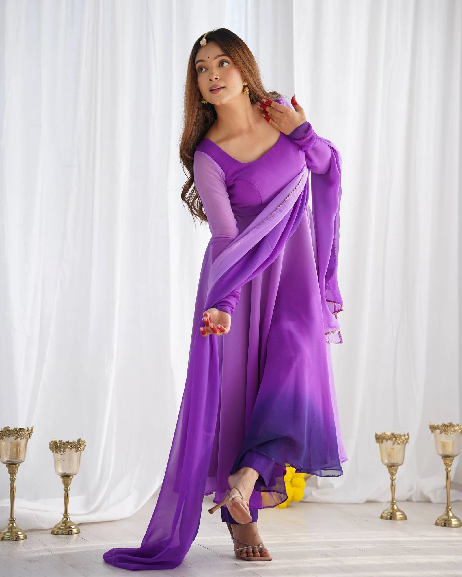 Woman in a long purple dress standing in front of white curtains with gold candle holders on either side.