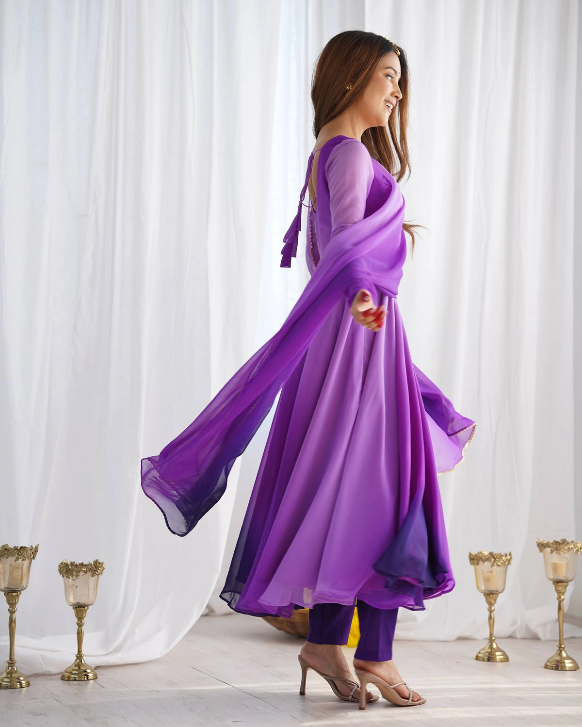 A profile shot of a woman in a purple Anarkali suit looking over her shoulder with a smile, showing the back detail and the flow of the dress.