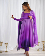 A woman stands in a bright room wearing a violet and purple ombre Anarkali suit, holding a matching sheer dupatta draped across her arms.
