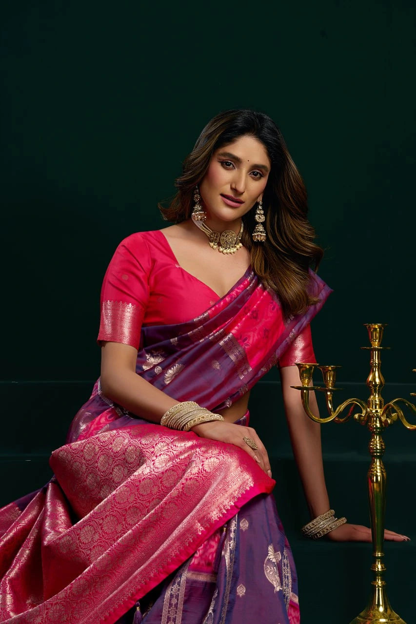 Woman in a pink and purple saree sitting next to a gold candle holder on a dark background