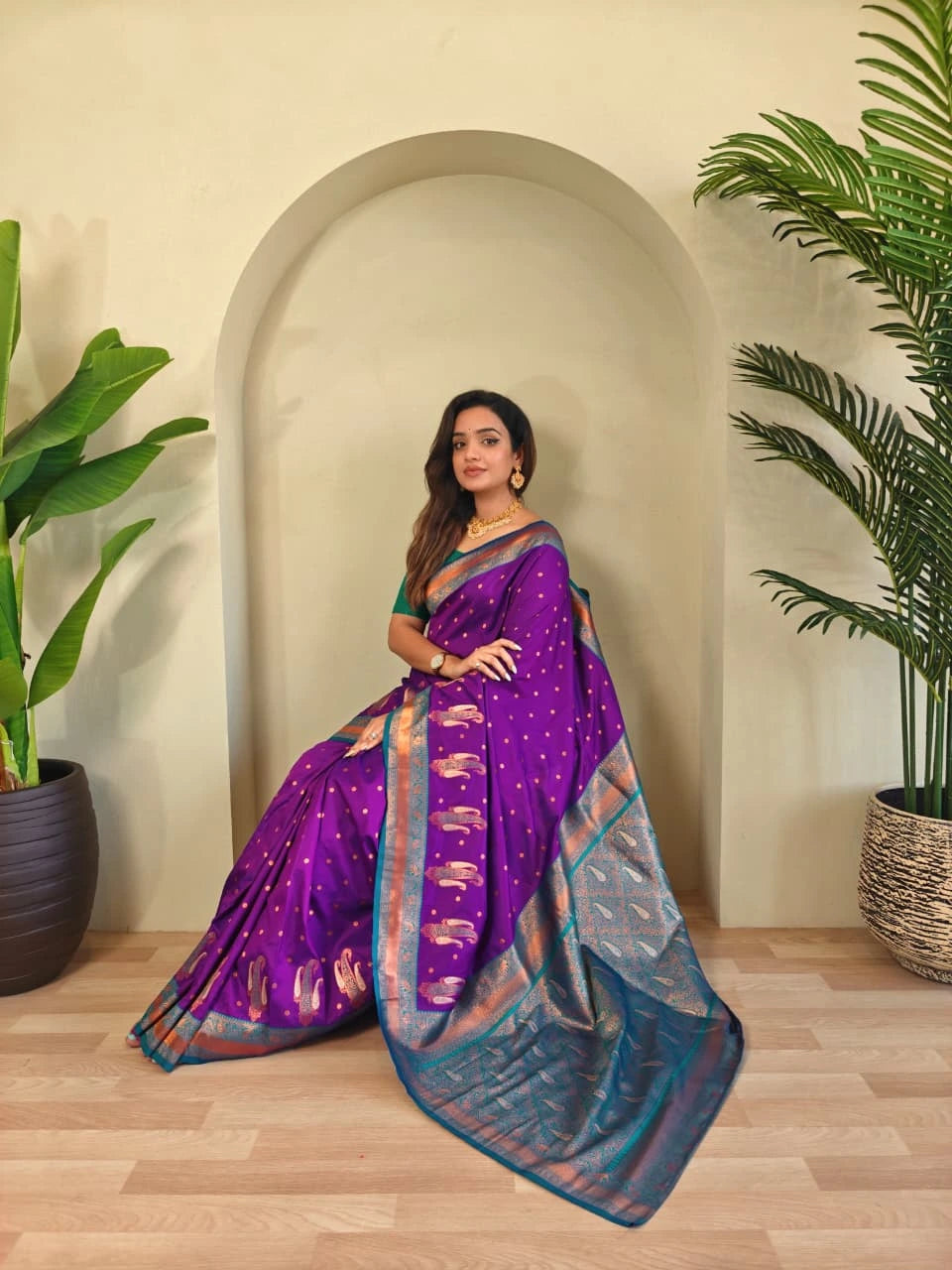 Woman in a purple saree standing indoors with plants on either side.