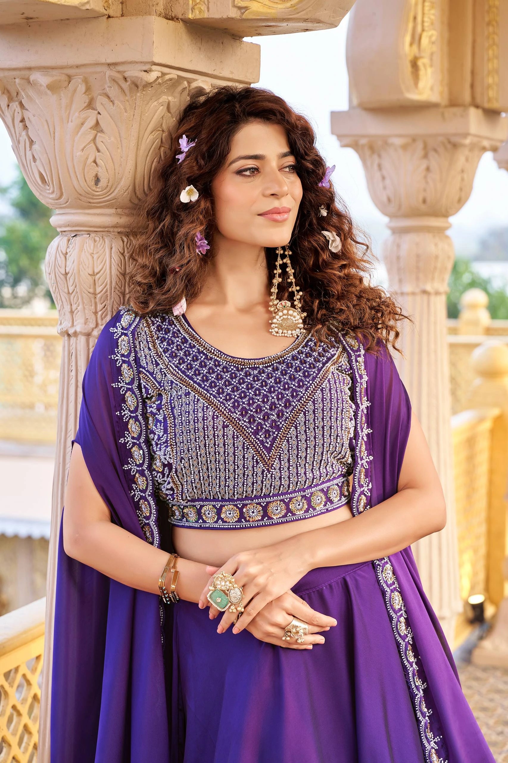 Woman in a purple traditional outfit with intricate designs standing in front of decorative columns.