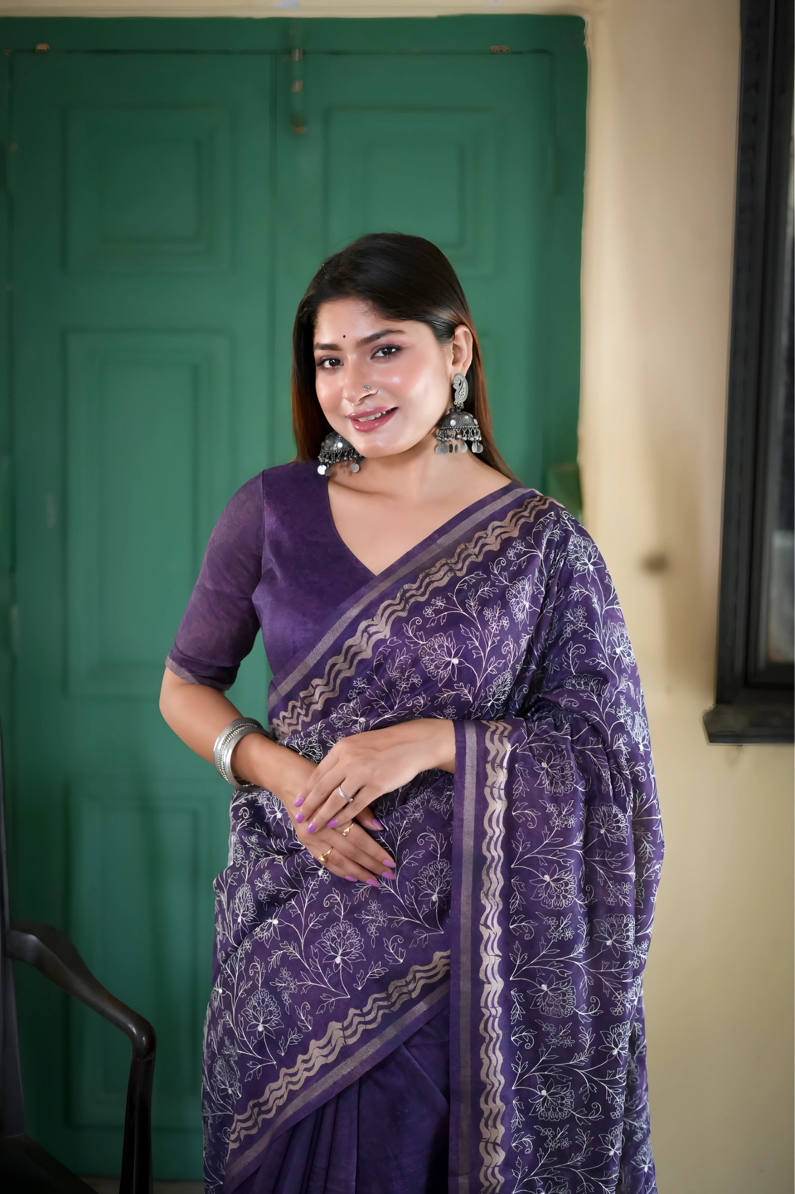 A woman models an elegant Deep Purple Tussar Silk Saree. The saree features dense all-over floral embroidery/weaving in a light metallic thread, framed by a contrasting metallic border. She wears a matching purple blouse and accessorizes with heavy oxidized silver jhumka earrings and bangles, posing indoors next to a teal door.