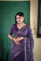 A woman models an elegant Deep Purple Tussar Silk Saree. The saree features dense all-over floral embroidery/weaving in a light metallic thread, framed by a contrasting metallic border. She wears a matching purple blouse and accessorizes with heavy oxidized silver jhumka earrings and bangles, posing indoors next to a teal door.