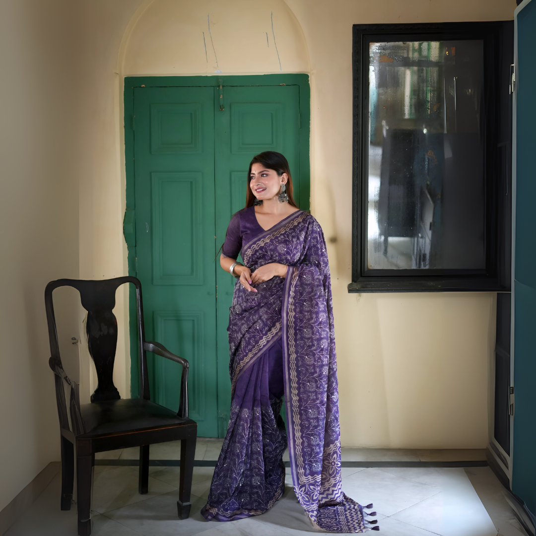 A woman models an elegant Deep Purple Tussar Silk Saree. The saree features dense all-over floral embroidery/weaving in a light metallic thread, framed by a contrasting metallic border. She wears a matching purple blouse and accessorizes with heavy oxidized silver jhumka earrings and bangles, posing indoors next to a teal door.
