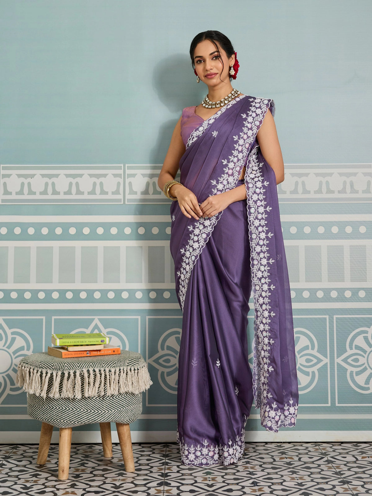 Woman in a purple saree with white patterns standing in a room with decorative wall and stool.