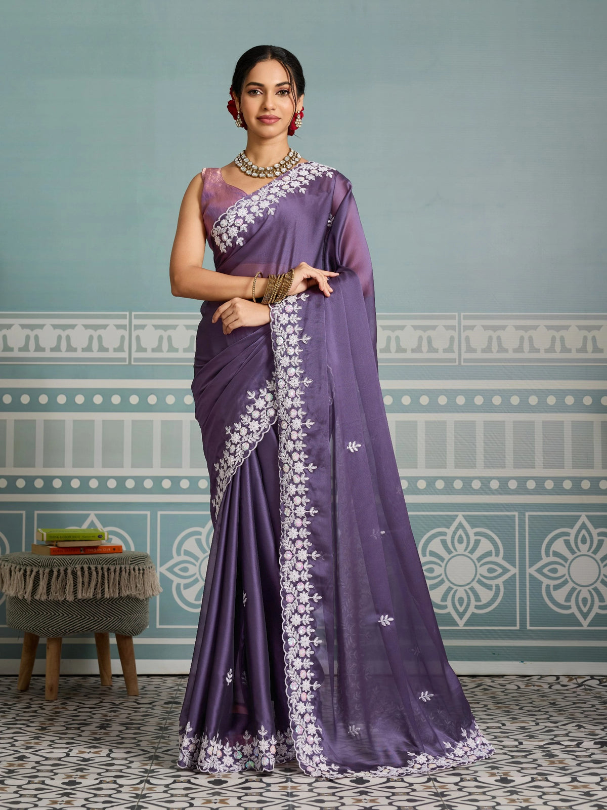 Woman wearing a purple saree with white embroidery against a decorative wall.