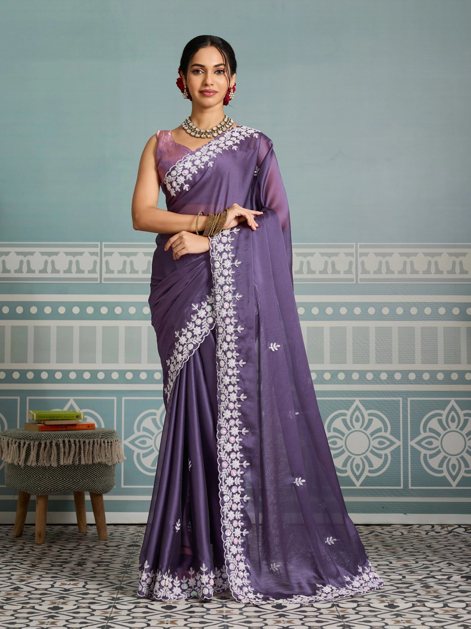 Woman wearing a purple saree with white embroidery against a decorative wall.