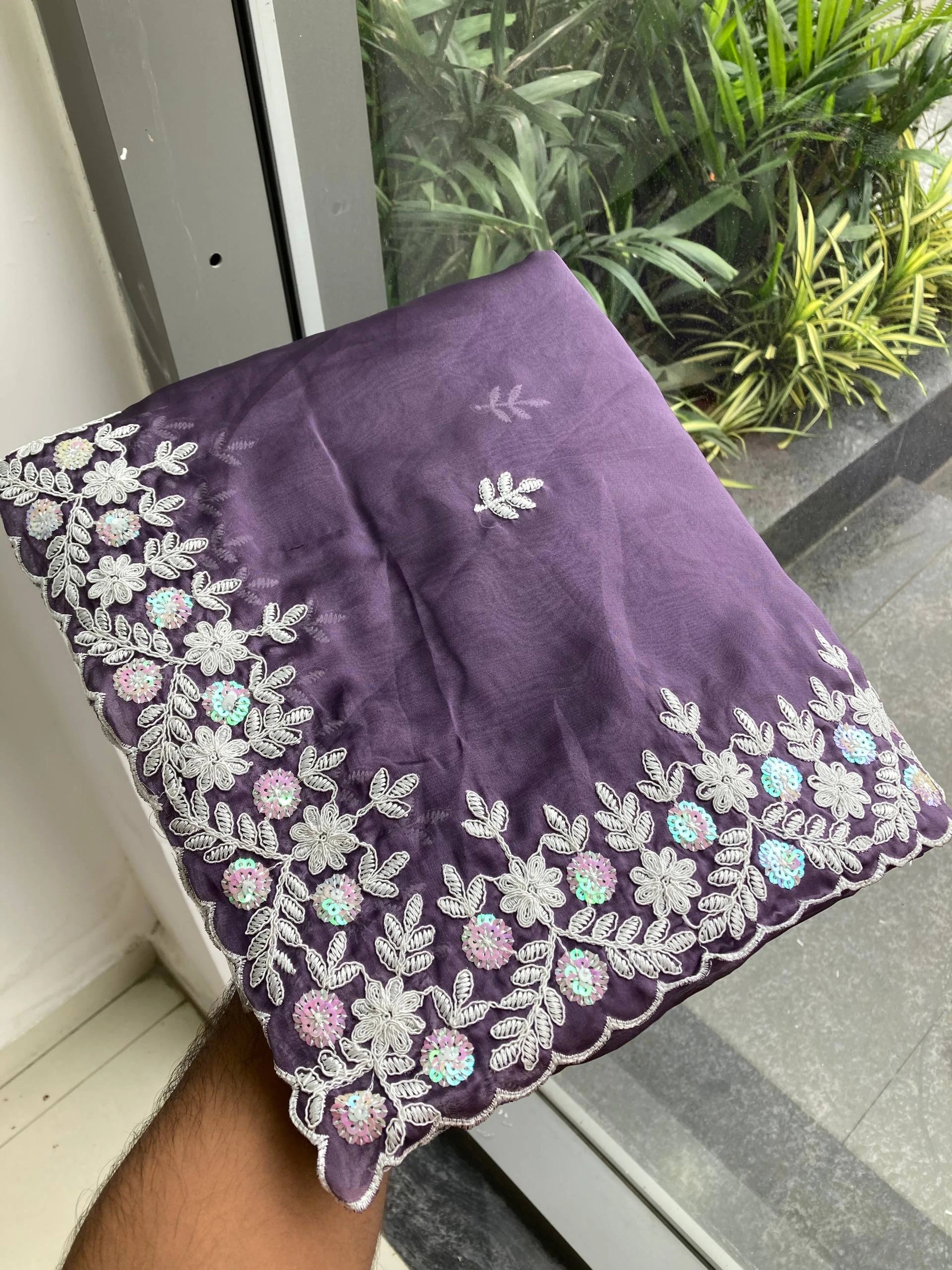 Purple fabric with floral embroidery held by a hand against a glass door with plants in the background