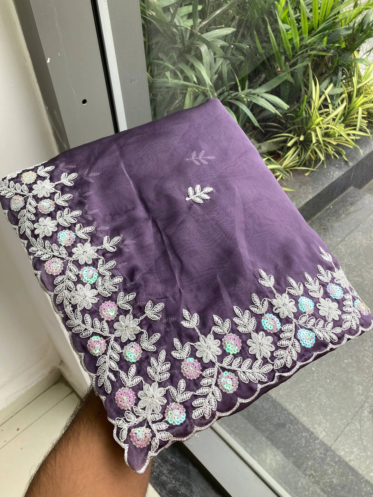 Purple fabric with floral embroidery held by a hand against a glass door with plants in the background