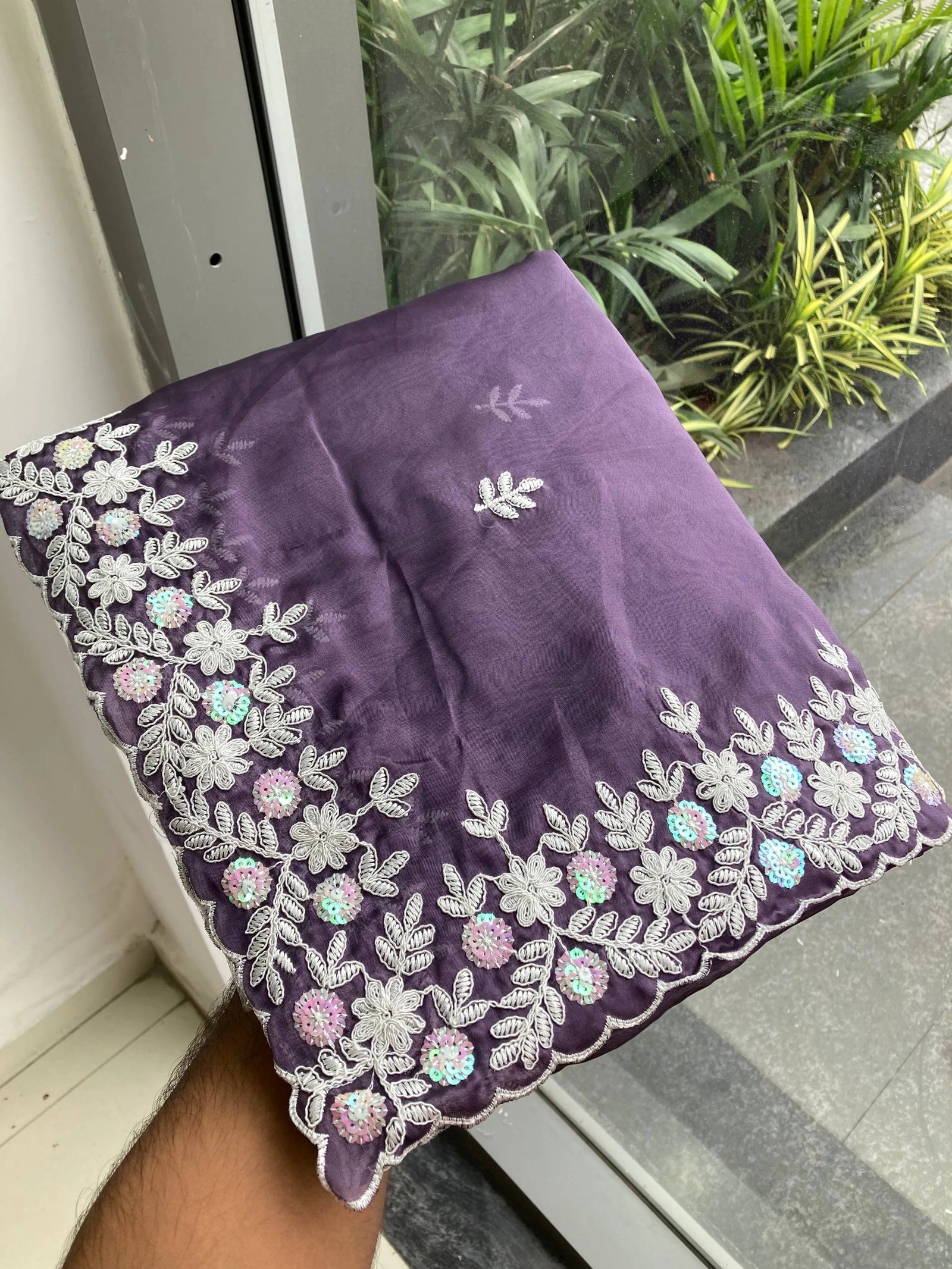 Purple fabric with floral embroidery held by a hand against a glass door with plants in the background
