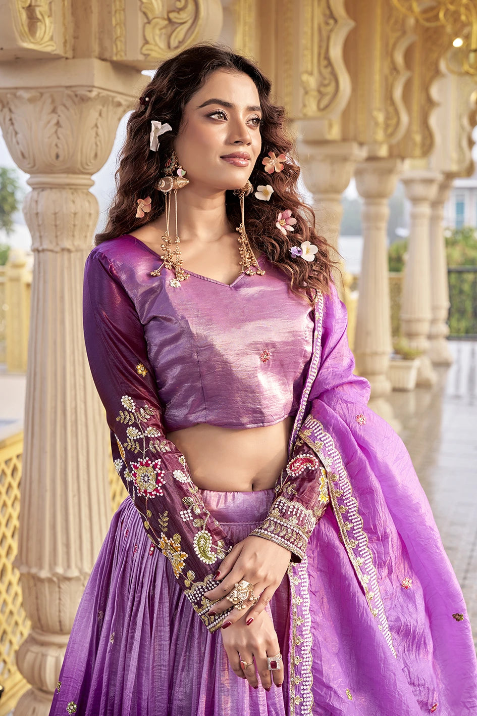 Woman in a traditional purple outfit with floral details standing in front of decorative columns.