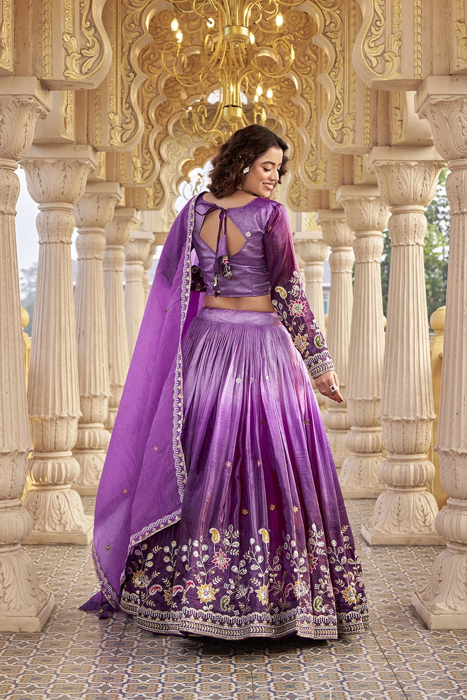 Woman in a purple embroidered traditional outfit standing in an ornate architectural setting.