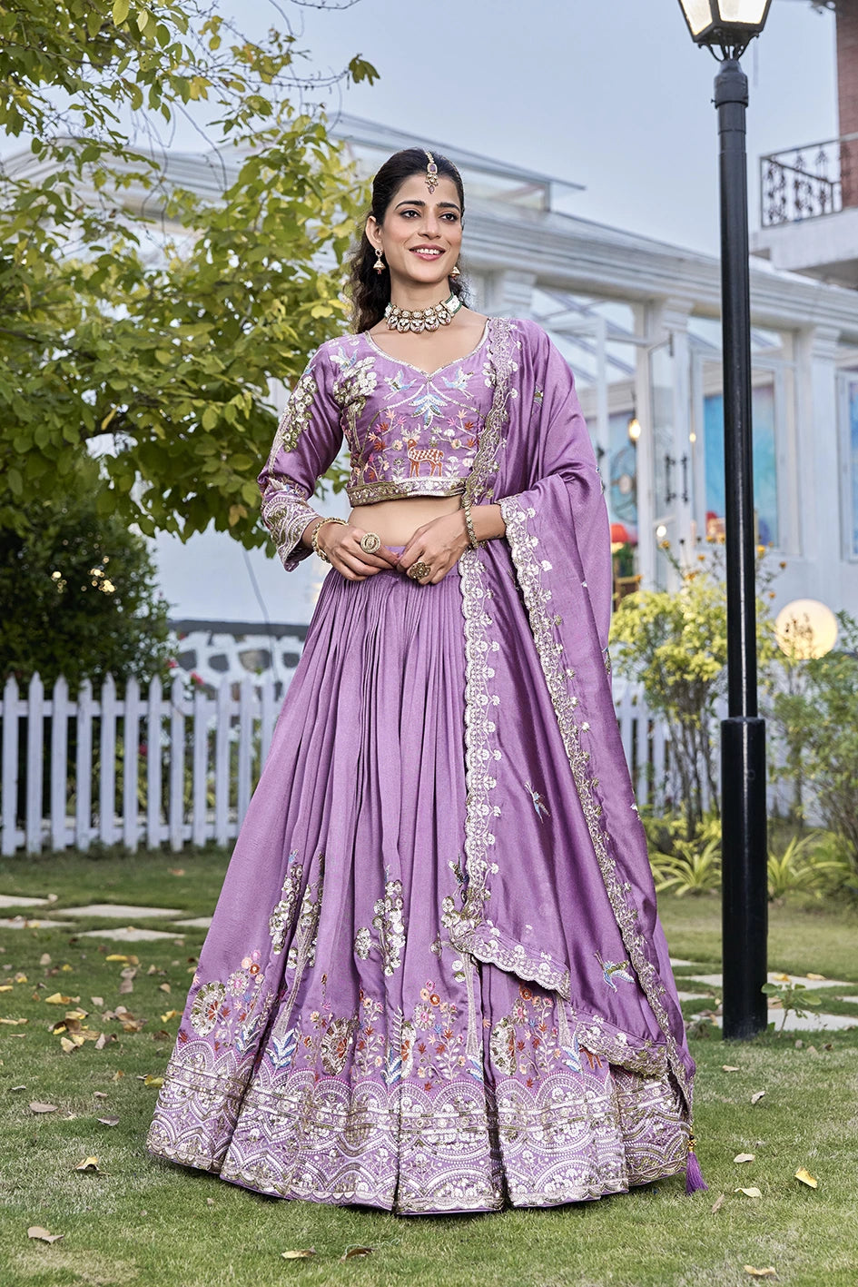 Woman in a purple traditional outfit standing outdoors with greenery and a building in the background