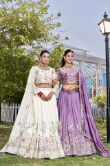 Two women in traditional embroidered lehengas standing outdoors with trees and a building in the background.