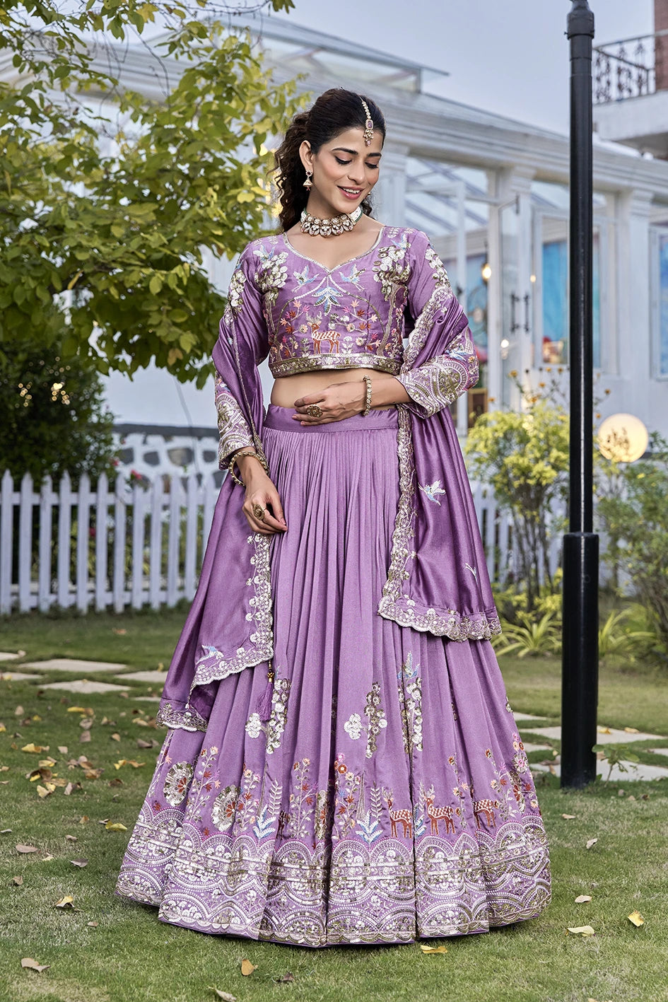 Woman in a purple embroidered traditional outfit standing outdoors with greenery.