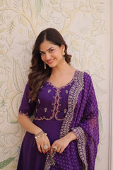 Woman wearing a purple traditional outfit with intricate designs against a decorative wall.
