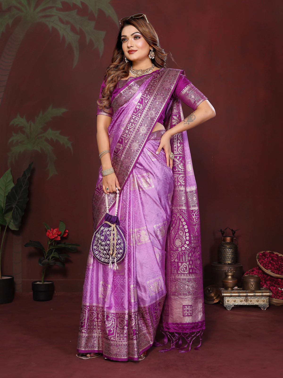 Woman wearing a purple saree with a matching blouse, holding a handbag, against a dark background.