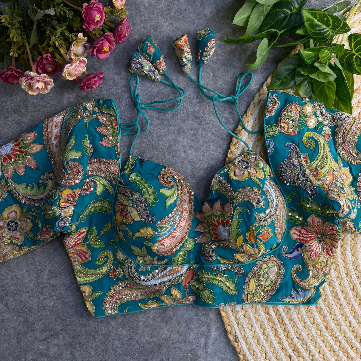 Teal padded blouse with intricate multicolor floral and paisley embroidery, sequin accents, and tie-back detail, displayed on a grey surface with artificial flowers and green leaves in the background.