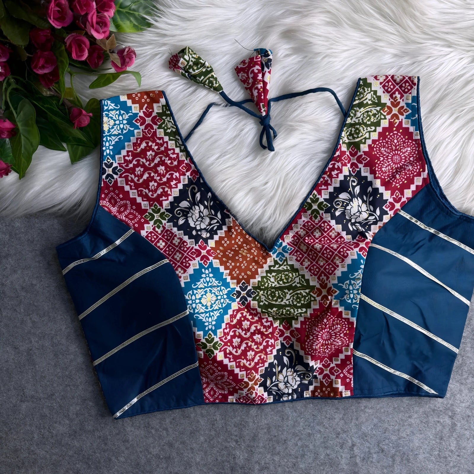 Traditional multicolor Ajrak-print blouse featuring striking geometric and floral motifs in navy blue, red, green, orange, and light blue. The blouse has a deep V-neckline, navy silk side panels with metallic silver stripes, and a matching tasseled tie-back, displayed flat on a white fur background with artificial pink flowers in the corner.