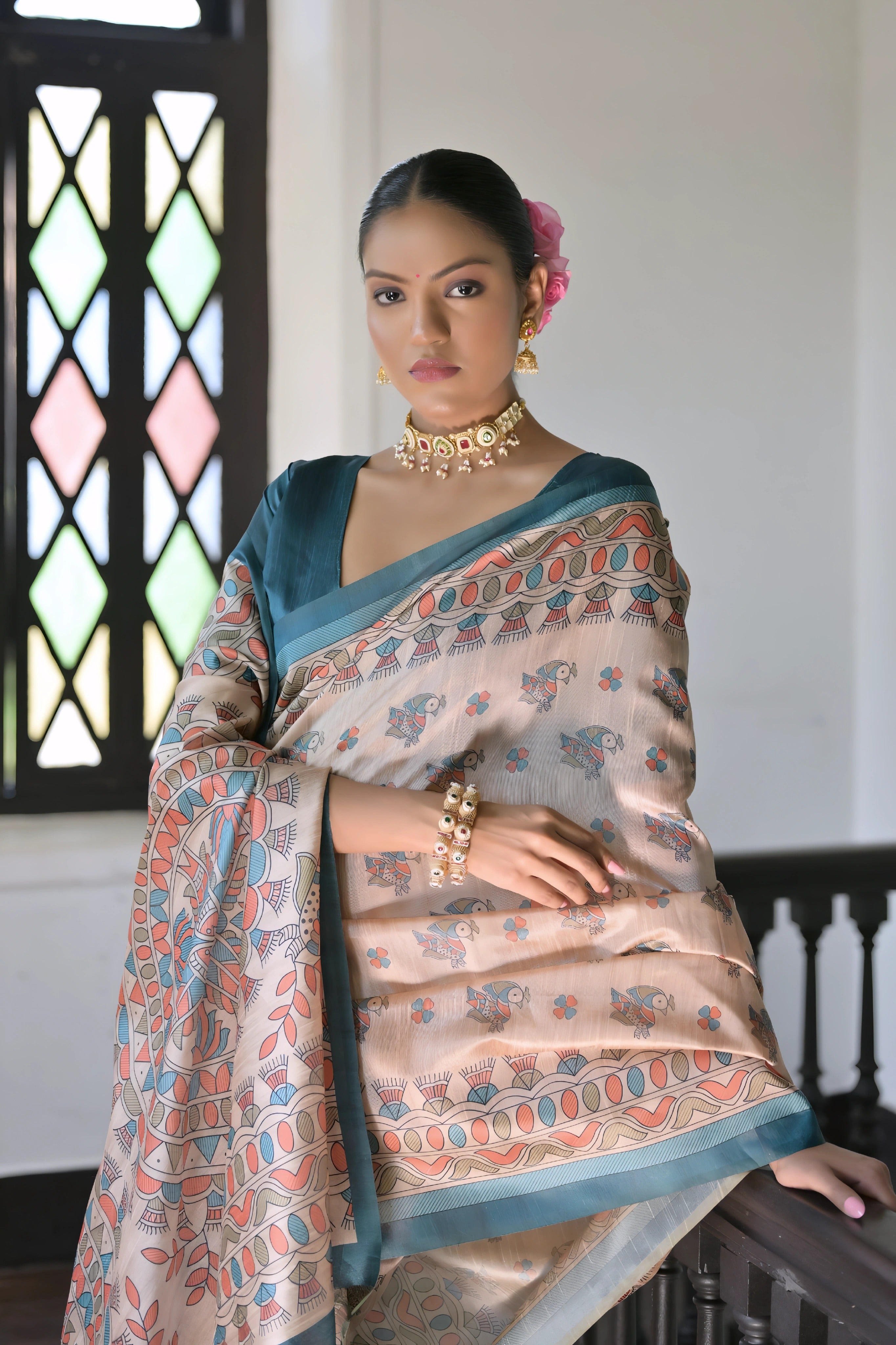 A woman models an elegant Beige Soft Tussar Silk Saree featuring an all-over Madhubani-style print in teal and coral/rust. The saree is framed by a solid Rama Blue border. She wears a deep teal short-sleeve blouse and a gold and pearl choker necklace, posing indoors next to a dark wooden banister.