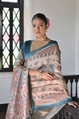 A woman models an elegant Beige Soft Tussar Silk Saree featuring an all-over Madhubani-style print in teal and coral/rust. The saree is framed by a solid Rama Blue border. She wears a deep teal short-sleeve blouse and a gold and pearl choker necklace, posing indoors next to a dark wooden banister.