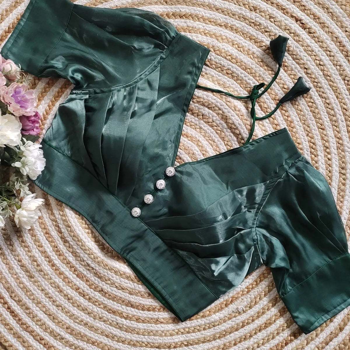 Rama Blue satin blouse with pleated detailing and four decorative silver buttons on the front, featuring elbow-length sleeves and tie-up tassels, displayed on a circular woven mat with white and pink flowers on the side.