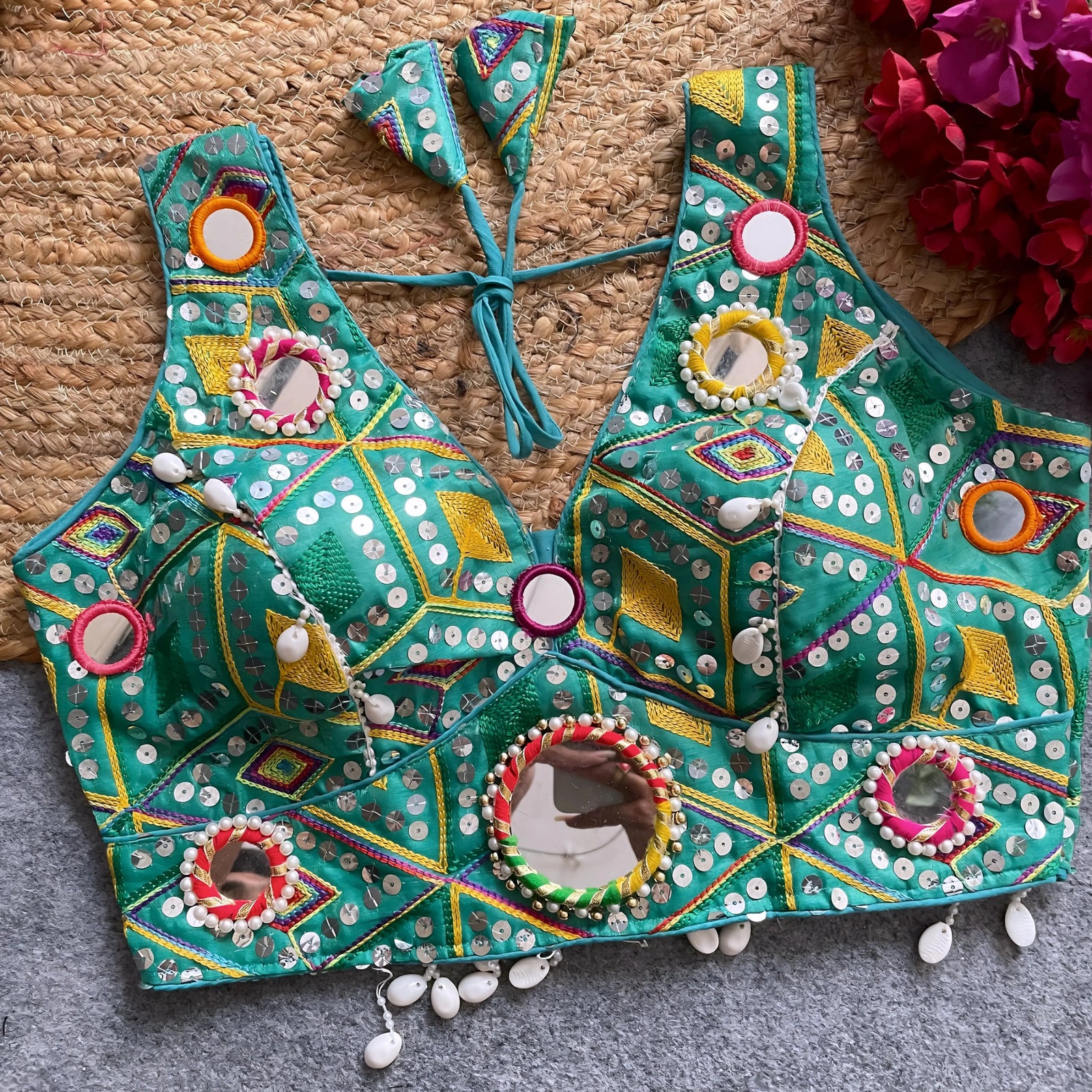 raditional teal Gujarati-style blouse featuring vibrant multicolor geometric embroidery, round mirrors bordered with colorful beads, and intricate white cowrie shell accents along the hem. The blouse includes tasseled tie-back detail and is displayed flat on a grey carpet with a woven mat and fuchsia artificial flowers in the corner.