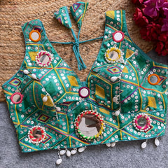 raditional teal Gujarati-style blouse featuring vibrant multicolor geometric embroidery, round mirrors bordered with colorful beads, and intricate white cowrie shell accents along the hem. The blouse includes tasseled tie-back detail and is displayed flat on a grey carpet with a woven mat and fuchsia artificial flowers in the corner.