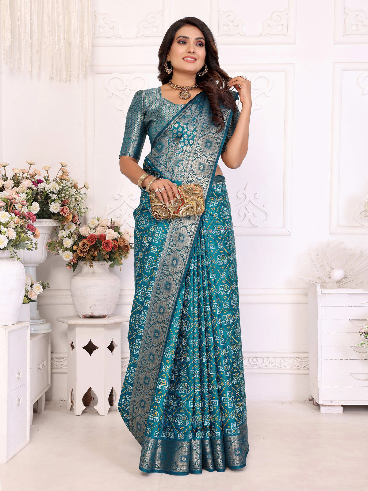 Woman in a blue saree holding a clutch in a decorated room with flowers and furniture.