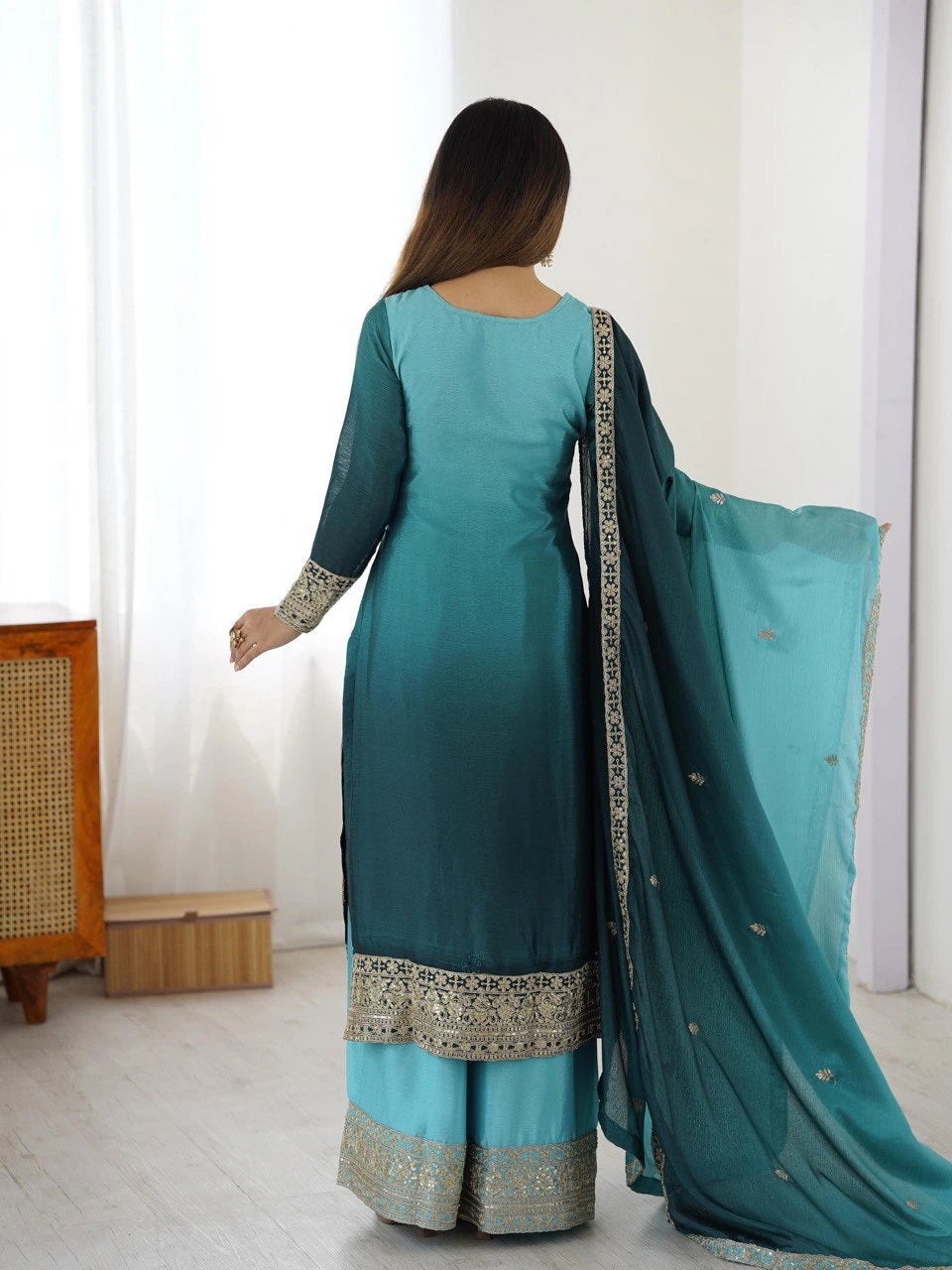 Woman wearing a teal traditional outfit with a matching dupatta in a minimal indoor setting.