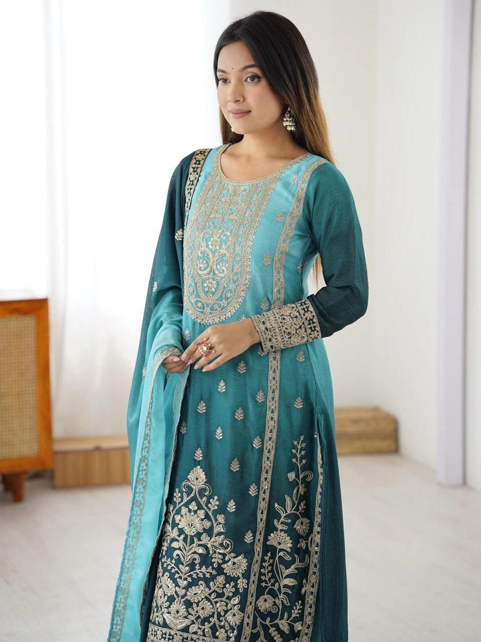 Woman wearing a teal traditional outfit with intricate silver embroidery in a minimal indoor setting.